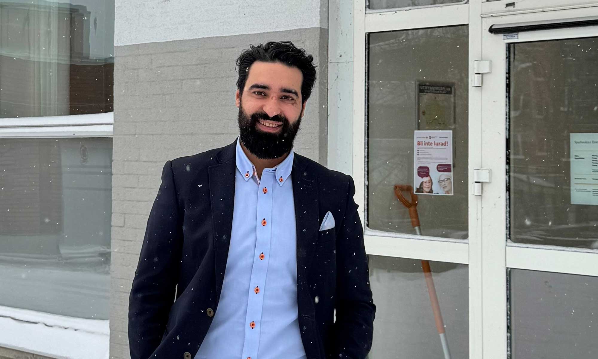 a man in a dark jacket and blue shirt standing outside, it is snowing